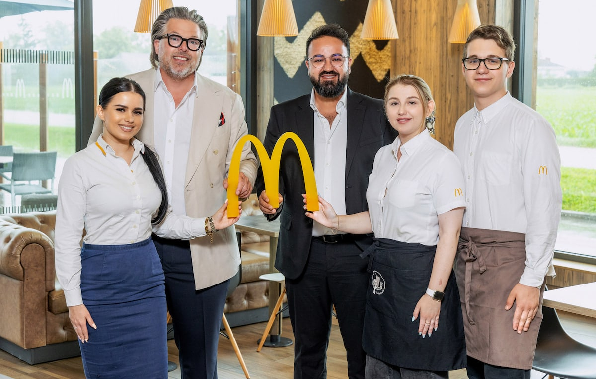Sosdar Abdalkarim und Michael Kühlwein halten zusammen mit drei McDonald’s-Mitarbeiter:innen das McDonald’s-Logo in der Hand. Im Hintergrund ist der Innenraum des McDonald’s-Restaurants in Eferding zu sehen.