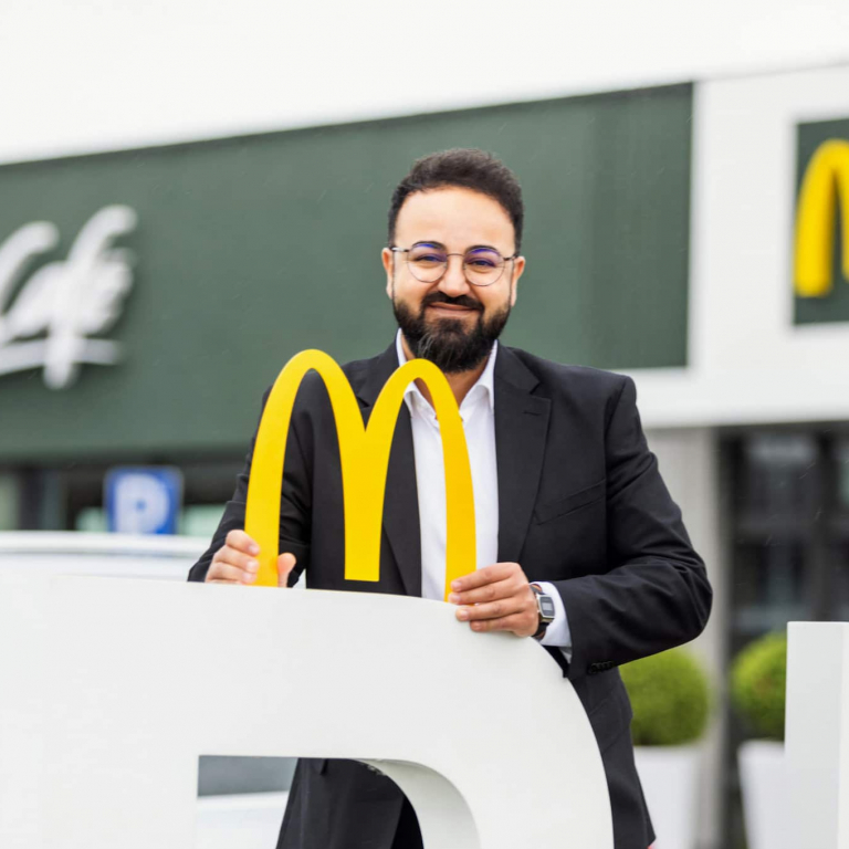 Sosdar Abdalkarim lächelt und hält das McDonald’s-Logo in der Hand. Im Hintergrund ist die McDonald’s-Filiale in Eferding zu sehen.